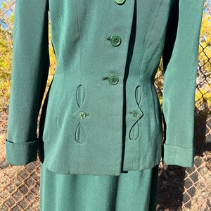 May include: A dark green suit featuring a tailored jacket with decorative buttons and a matching skirt. The jacket has a fitted silhouette with unique button detailing and a classic collar. The fabric appears to be a smooth, solid color, suitable for formal or professional wear.