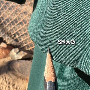 May include: Close-up of a dark green fabric with the word "SNAG" printed in white. A sharpened pencil points towards a small imperfection in the fabric. The fabric has a textured appearance.