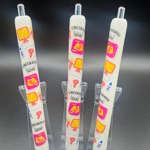 Peut inclure: Trois stylos à paillettes blanches avec des pointes d'encre noires. Chaque stylo est décoré d'un motif répété d'une image de sonogramme, d'un cœur rose et des mots "SONOGRAPHY SQUAD". Les stylos sont exposés sur des présentoirs en plastique transparent.