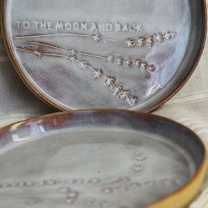 May include: Two ceramic plates with a light grey glaze and a brown rim. The top plate has the text "To the moon and back" and a design of lavender flowers. The bottom plate has a similar design of lavender flowers.
