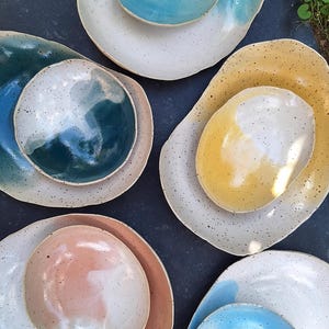 May include: A collection of handmade ceramic plates and bowls in various sizes and colors. The plates feature speckled white backgrounds with colorful accents in shades of blue, yellow, pink, and green. The bowls are nested inside the plates.