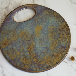 May include: A round ceramic serving tray with a floral design in shades of blue, green, and brown. The tray has a handle cut out in the center.