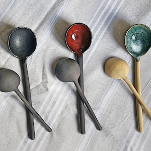 May include: Five ceramic spoons in different colors, including black, red, green, and beige. The spoons are arranged on a white and gray striped fabric.