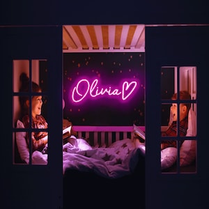 May include: Two children are reading in a bunk bed with a pink neon sign that says "Olivia" with a heart. The sign is on a dark wall with gold stars.