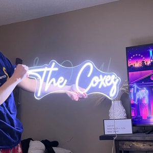 Puede incluir: Un letrero de ne&oacute;n blanco con el texto "The Coxes" en una fuente cursiva. El letrero est&aacute; siendo sostenido por la mano de una persona.