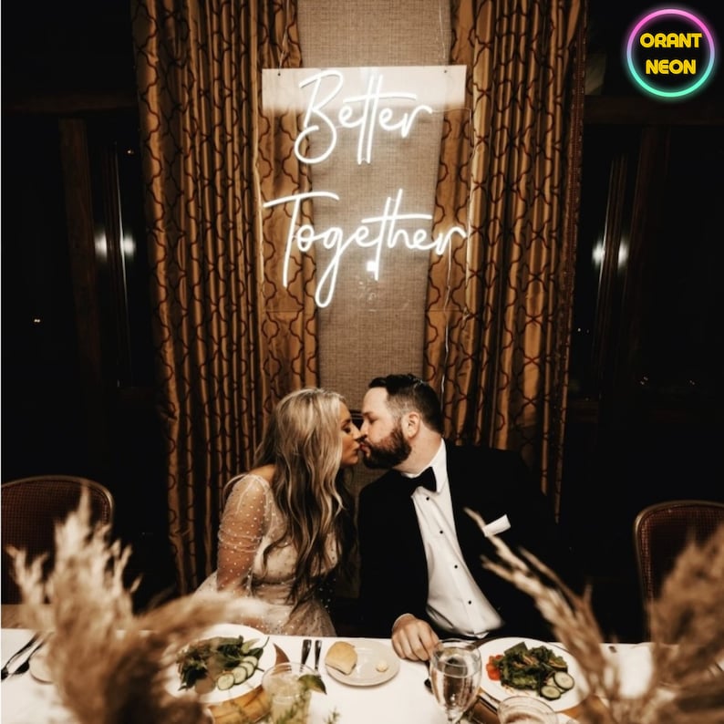 May include: A couple kisses in front of a white neon sign that reads "Better Together". The sign is hanging on a wall with gold patterned curtains. The couple is sitting at a table with white linens and a centerpiece of dried grasses.