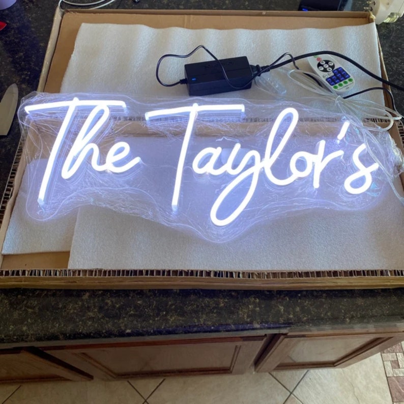 Puede incluir: Un letrero de ne&oacute;n blanco con el texto "The Taylor's" en una fuente cursiva. El letrero est&aacute; empaquetado en una caja de cart&oacute;n con cacahuetes de embalaje.
