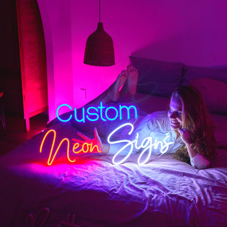 Puede incluir: Una mujer est&aacute; tumbada en una cama con un letrero de ne&oacute;n que dice "Custom Neon Signs". El letrero est&aacute; formado por tres piezas separadas, cada una con un ne&oacute;n de color diferente. El letrero est&aacute; encendido y brilla intensamente.