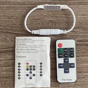 May include: A white LED controller with a remote control. The controller has a white wire loop and a connector. The remote control has buttons for light, mode, and speed. The text on the controller reads "LED CONTROLLER with RF Remote Control Peak Output Current 15A". The text on the remote control reads "RF Wireless".