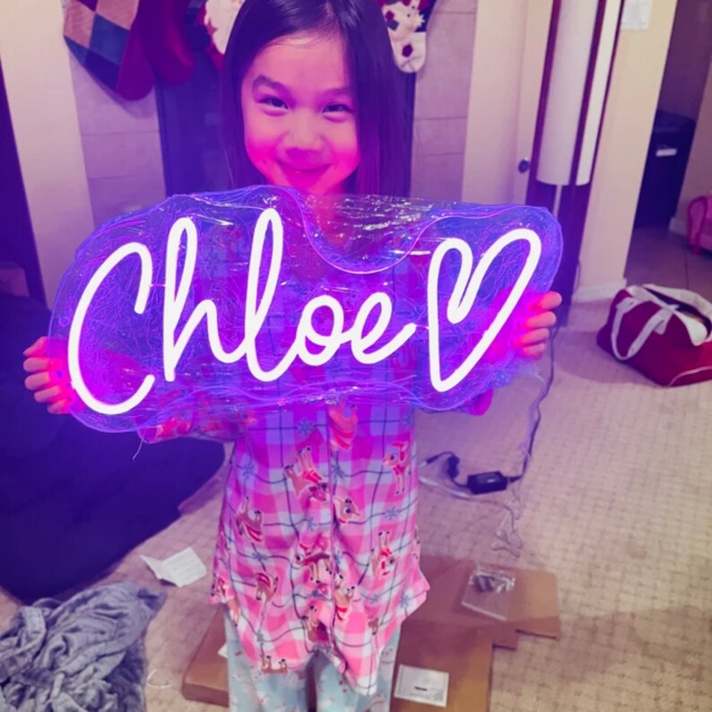 Puede incluir: Una ni&ntilde;a sostiene un letrero de ne&oacute;n morado y blanco con el nombre "Chloe" y un coraz&oacute;n. El letrero est&aacute; iluminado y tiene un contorno blanco.