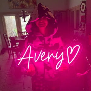 Puede incluir: Un letrero de ne&oacute;n rosa con el nombre "Avery" y un coraz&oacute;n. El letrero lo sostiene una persona que lleva una sudadera con capucha de estampado de vaca en blanco y negro.