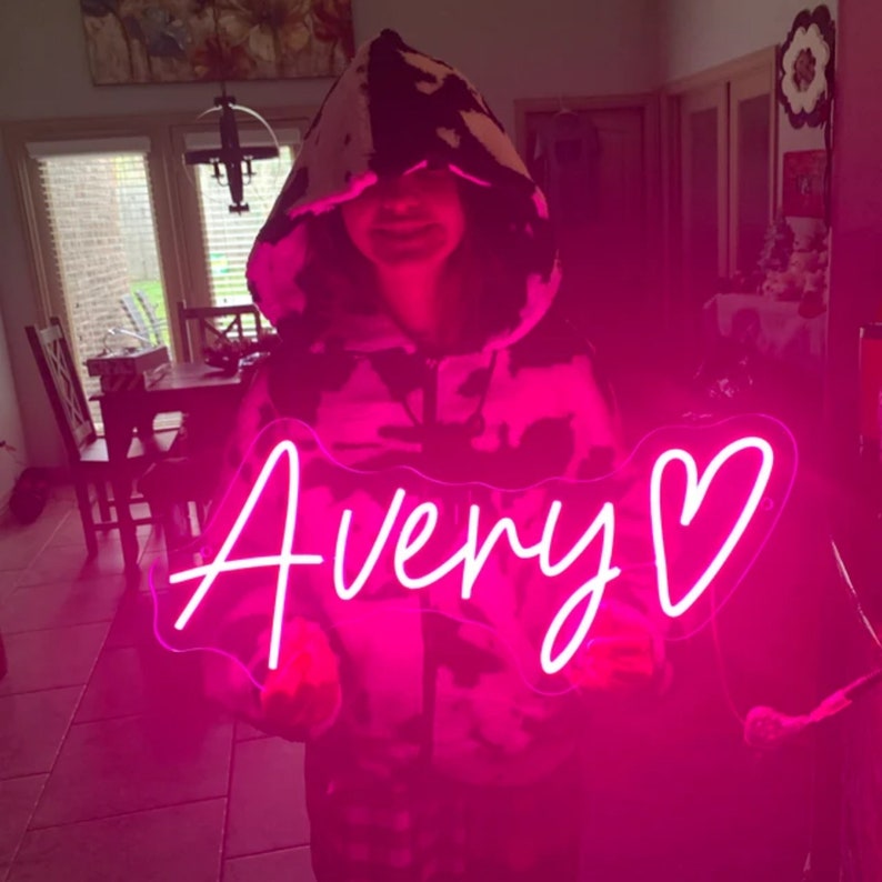 Puede incluir: Un letrero de ne&oacute;n rosa con el nombre "Avery" y un coraz&oacute;n. El letrero lo sostiene una persona que lleva una sudadera con capucha de estampado de vaca en blanco y negro.