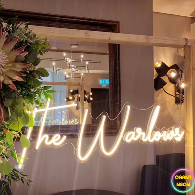 May include: A white neon sign with the text "The Warlows" in a cursive font. The sign is hanging from a wooden frame and is illuminated against a dark background.