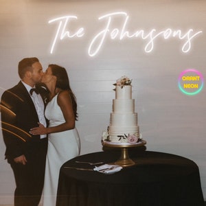 May include: A couple kisses in front of a white wall with a neon sign that reads "The Johnsons". A white wedding cake with pink flowers sits on a gold stand on a black table.