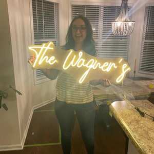 May include: A woman holds up a custom neon sign that reads "The Wagner's". The sign is lit up and has a white neon glow. The sign is made of a clear acrylic material and has a white backing.