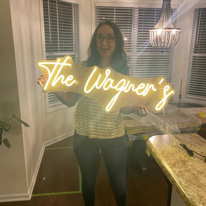 May include: A woman holds up a custom neon sign that reads "The Wagner's". The sign is lit up and has a white neon glow. The sign is made of a clear acrylic material and has a white backing.