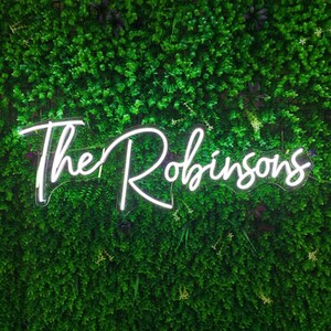 Puede incluir: Un letrero de ne&oacute;n blanco con el texto "The Robinsons" en una pared verde de follaje artificial.