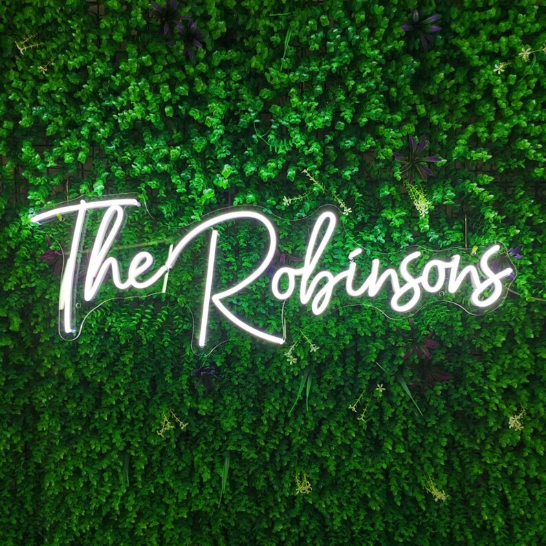 Puede incluir: Un letrero de ne&oacute;n blanco con el texto "The Robinsons" en una pared verde de follaje artificial.
