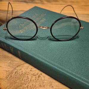 May include: Vintage round eyeglasses with dark brown frames and thin wire temples rest on a green book. The book has gold lettering that reads "Sunday School Lessons". The glasses have a gold bridge.