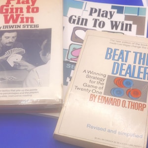 May include: A stack of vintage books with titles related to card games. The books include "Play Gin to Win" and "Beat the Dealer." The covers feature text and illustrations, with a blue background.