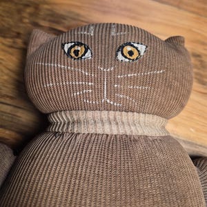 May include: A brown stuffed animal shaped like a cat, crafted from ribbed knit fabric. The face features embroidered yellow eyes with white accents and white stitched whiskers. A ribbed collar separates the round head from the body.