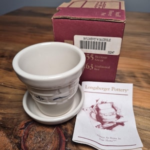 May include: A small off-white ceramic flower pot with a matching saucer, featuring a blue painted floral design. The set includes a maroon cardboard box labeled SM FLOWER POT W/ SAUCER BLUE and a Longaberger Pottery pamphlet with a pitcher illustration.