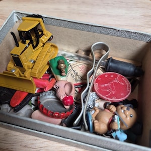 May include: A rectangular cardboard box containing a collection of small items including a yellow toy bulldozer, a pink pig figurine, a red circular token with a deer illustration, a silver metal hair clip, and a small brown bear figurine.