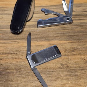 May include: A black leather case and two multi-tool pocket knives with various tools, including a knife blade, nail file, and bottle opener. The tools are made of shiny metal and are open on a wooden surface.