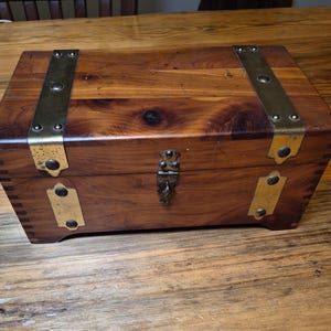 May include: A rectangular wooden box with brass accents. The box is made of a reddish-brown wood and features brass corner protectors and a clasp. The box is sitting on a wooden surface.