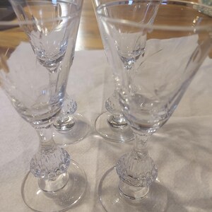 May include: Four clear, stemmed glasses with etched floral designs. Each glass has a round base, a decorative bulbous stem, and a flared bowl. The glasses are arranged on a white surface.