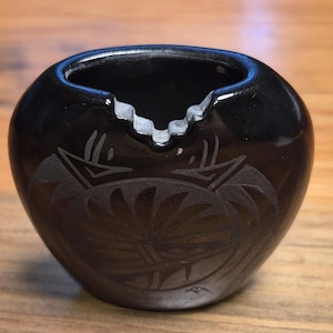 May include: A small, polished black ceramic vessel with a rounded, bulbous shape. The rim features a distinct, stepped cutout design. The front surface is decorated with a matte, etched geometric pattern and a small signature mark near the base.