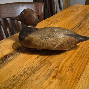 May include: A vintage wooden duck decoy with a brown head and a weathered, painted body in shades of gray and black. The duck is resting on a wooden table with a natural wood grain.