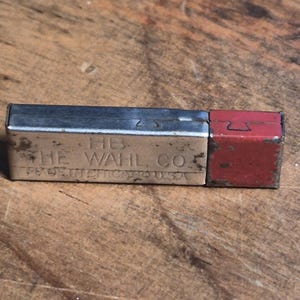 May include: A rectangular metal lead refill case with a silver-toned body and a red cap. The silver section is embossed with the text HB, THE WAHL CO, and MADE IN CHICAGO U.S.A. The metal surface shows signs of wear and minor oxidation throughout.