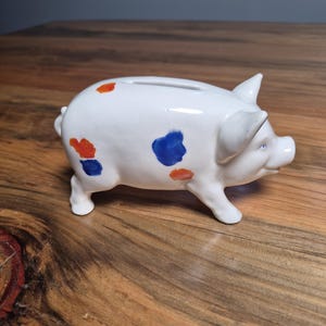 May include: A white ceramic piggy bank shaped like a pig with a slot on its back. The surface is decorated with scattered, hand-painted spots in vibrant shades of blue and orange. The pig features pointed ears, a snout, and a small curled tail.