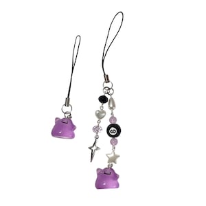 May include: Two phone charms featuring purple blob-shaped figures with simple facial expressions. One charm is a single figure on a black cord. The second charm includes a purple figure, a black eight-ball bead, white heart beads, and a silver star charm.