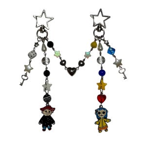 May include: Two silver keychains with star-shaped clasps. Each has a dangling design with beads, charms, and a character pendant. One keychain features a character in a black coat, the other in a yellow coat.
