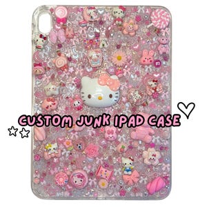May include: A clear iPad case decorated with pink and white embellishments. The case features Hello Kitty, flowers, bows, and candies. The text "CUSTOM JUNK IPAD CASE" is displayed on the case.