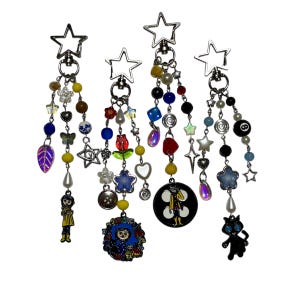 May include: Four beaded keychains with star-shaped clasps. Each keychain features a unique design with colorful beads, charms, and pendants. Charms include a girl in a yellow coat, a cat, and a black and white circle with a character.