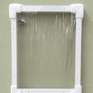May include: A white rectangular frame with protective corner padding and clear plastic sheeting. Small hardware is visible in a plastic bag attached to the frame. The frame is against a light green wall.