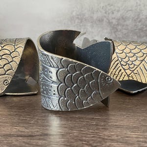 May include: Three metal napkin rings with fish designs. The rings are in shades of silver and gold, with detailed scales and fins. The fish are stylized and add a unique touch to any table setting.