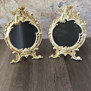May include: Two ornate, antique-style picture frames. Each frame has a cream-colored finish with gold accents and a cherub at the top. The frames have an oval opening with a black background and are set on a dark wooden surface.