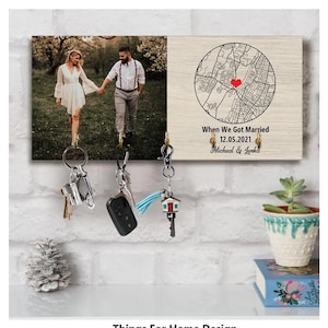 May include: A personalized wooden key holder featuring a photo and a map with a heart marking a location. The text reads "When We Got Married 12.05.2021 Micheal & Lenka." Several sets of keys hang from the hooks. The key holder is displayed against a white brick wall.