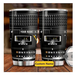 Op de afbeelding: Twee zwarte roestvrijstalen bekers met een camera lens ontwerp. De bekers hebben een zwart en zilver ontwerp met de tekst "YOUR NAME" en "Custom Name".