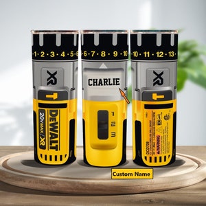 Könnte beinhalten: Drei gelbe und schwarze isolierte Becher mit einem Dewalt-Bohrer-Design. Der mittlere Becher trägt den Namen "CHARLIE". Die Becher stehen auf einer Holzoberfläche.