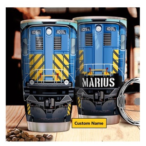 Peut inclure: Deux gobelets de voyage en acier inoxydable avec un motif de train bleu. Le train présente des rayures diagonales jaunes et le nom "MARIUS" en blanc. Les gobelets ont une étiquette "Nom personnalisé" et le numéro "105".