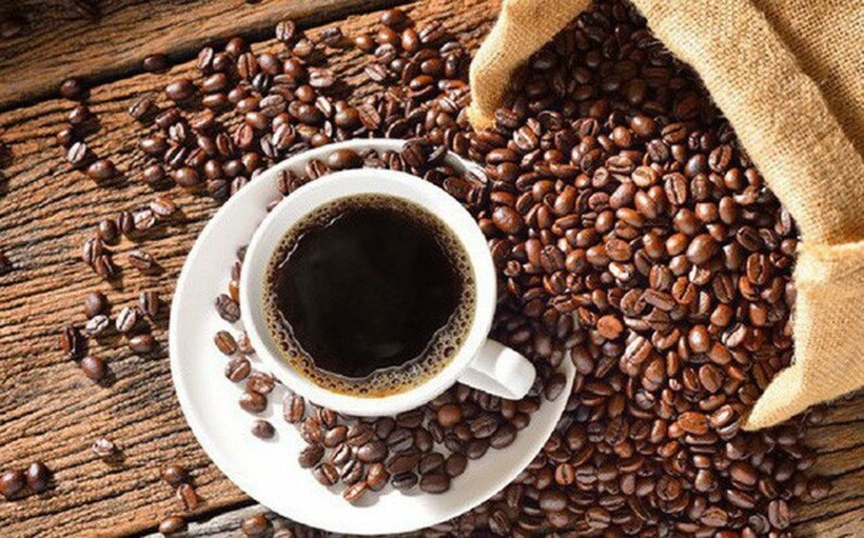May include: A white ceramic cup filled with black coffee sits on a white saucer surrounded by coffee beans. The cup has a handle and is partially filled with coffee. The coffee beans are scattered around the cup and saucer on a wooden surface.