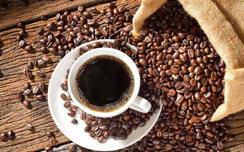 May include: A white coffee cup filled with dark coffee sits on a white saucer, surrounded by roasted coffee beans. A burlap sack spills more coffee beans onto a rustic wooden surface, creating a warm, inviting scene.