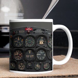 May include: A white ceramic mug with a black and white image of an aircraft cockpit instrument panel. The panel features various gauges and dials, including a climb indicator, a speed indicator, and a fuel gauge. The text "WARNING CRITICAL MACH NO.8" is visible on the panel.