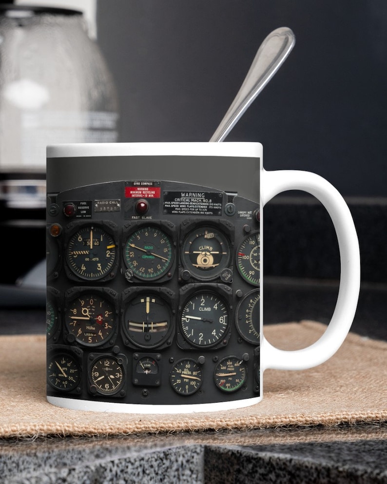 May include: A white ceramic mug with a black and white image of an aircraft cockpit instrument panel. The panel features various gauges and dials, including a climb indicator, a speed indicator, and a fuel gauge. The text "WARNING CRITICAL MACH NO.8" is visible on the panel.