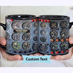 May include: A black ceramic mug with a design of an airplane cockpit instrument panel. The panel features various gauges and dials with numbers and labels. The mug has a handle and the text "NAME HERE U.S. AIRWAYS" is printed on the panel.